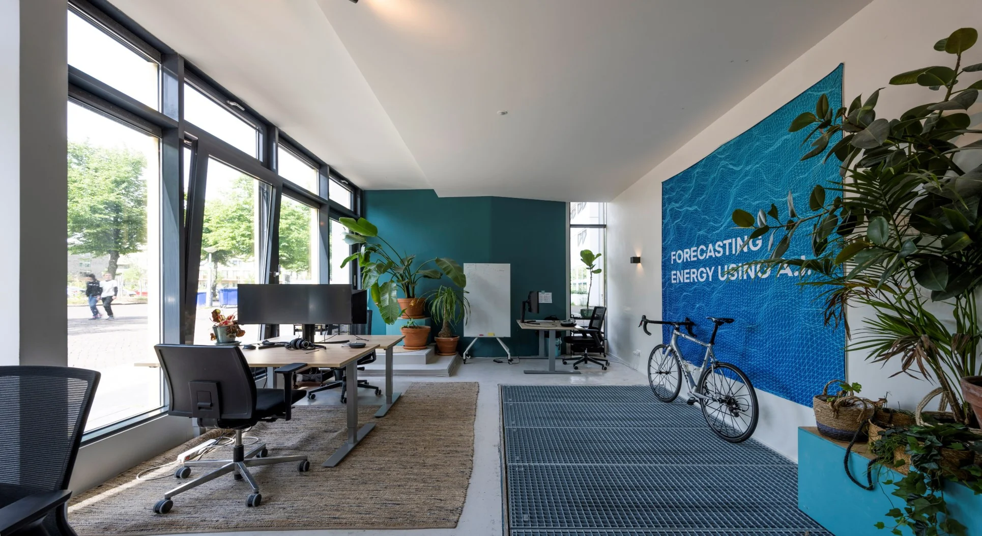 Moderne kantoorruimte aan de James Wattstraat met werkplekken, veel planten, een racefiets en een blauwe wand met de tekst "Forecasting Energy Using A.I.".
